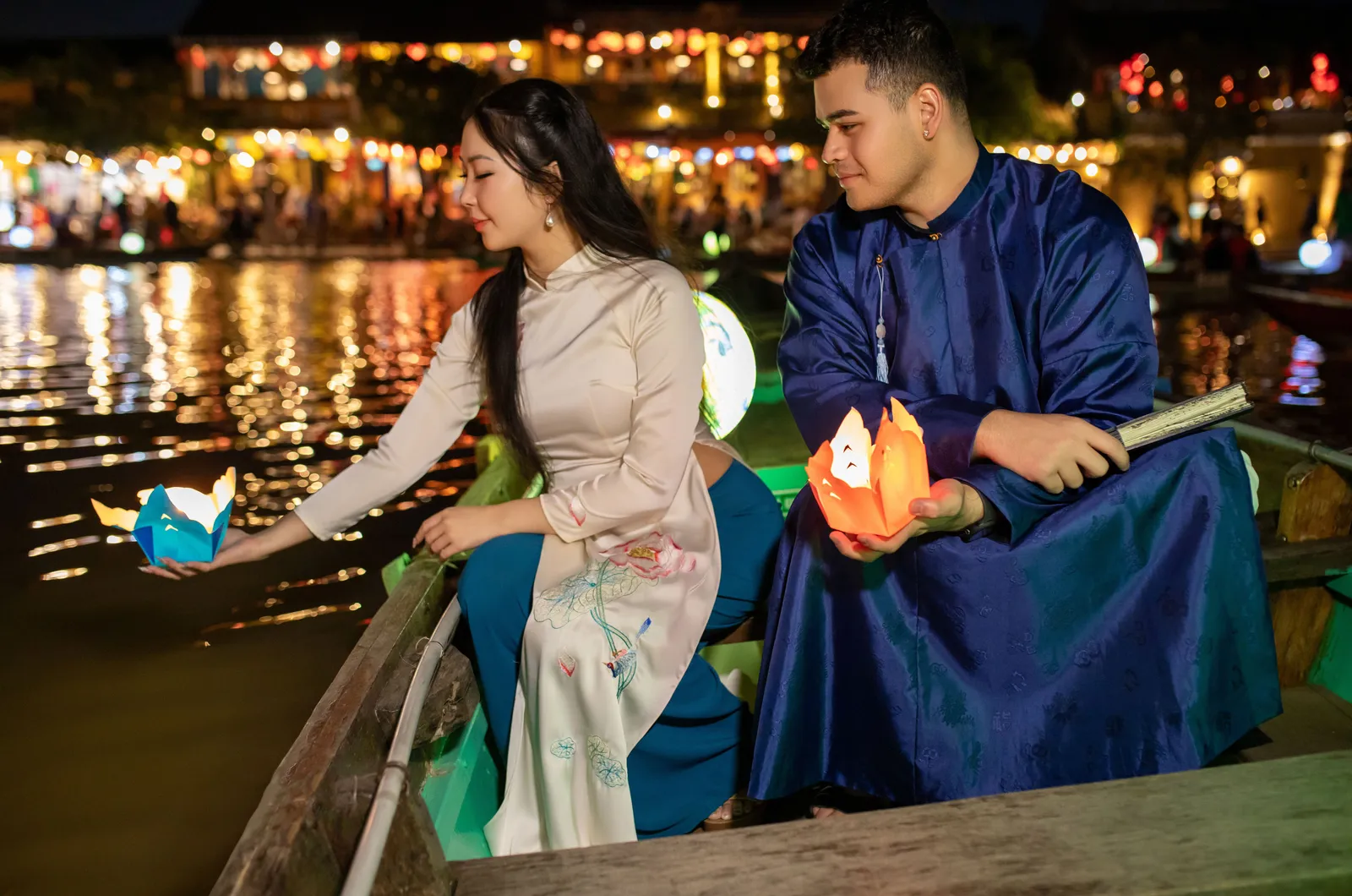 A couple in traditional ao dai releasing floating lanterns on the river at Hoi An, with the lantern lit old town behind