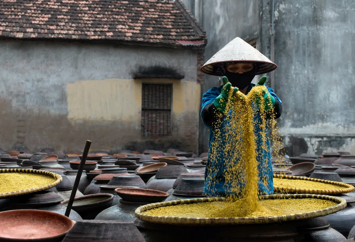 A woman in a conical hat working with grain beside clay jars in a village in central Vietnam