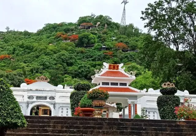 A white walled compound with a red tiled roof gate, a green hill behind