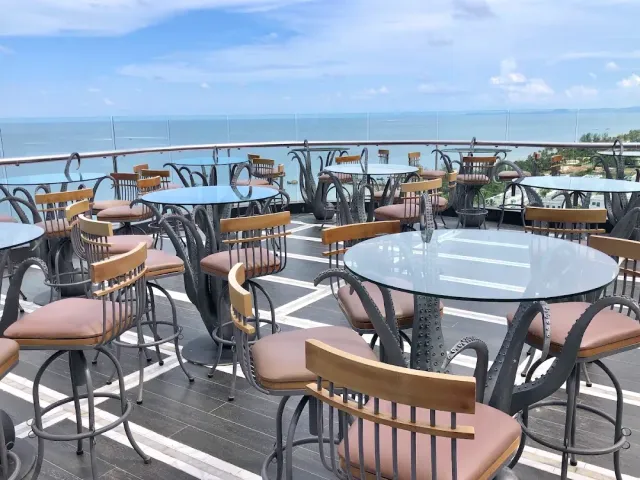 A rooftop terrace with round glass tables, ornate metal chairs and a clear railing facing the sea