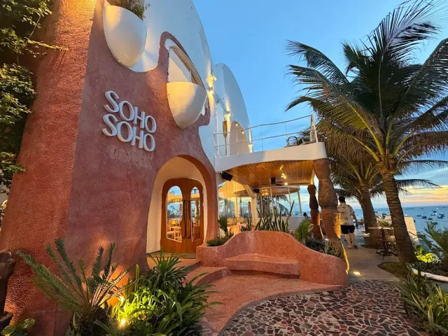 A sculptural pinkish building with rounded white forms, an arched wooden door and palms facing the sea