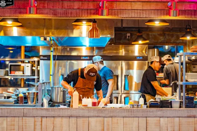 An open kitchen behind a counter, cooks in caps working under hanging copper lamps