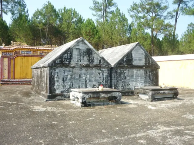 Two dark stone tomb structures with gabled tops and offering tables in front of them