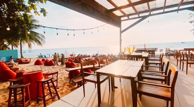 Red beanbags and wooden tables on the sand facing the sea at golden hour, with hanging lights