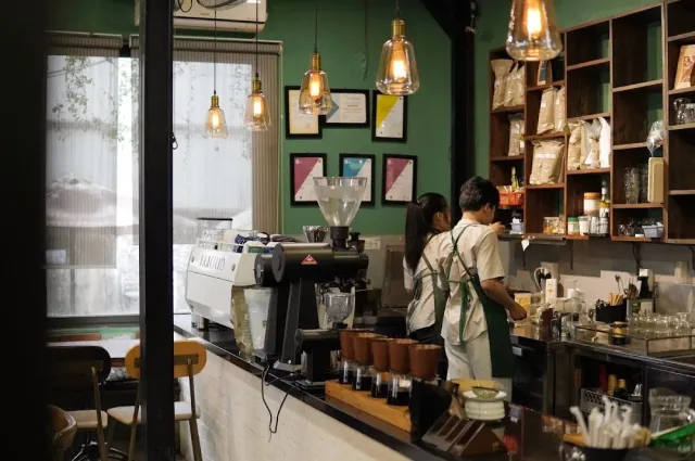 A preparation counter with an espresso machine and grinder, two baristas in green aprons and a green wall behind