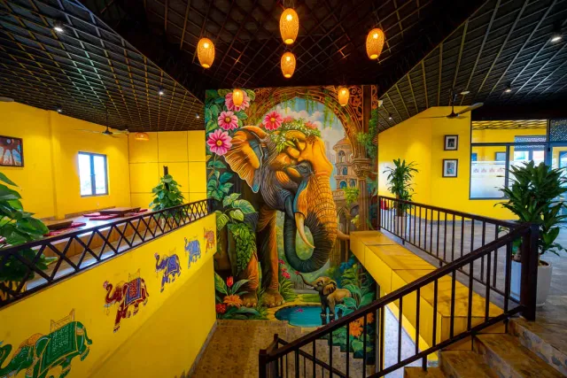 An indoor stairwell with yellow walls, a large painted elephant and orange lanterns on the ceiling