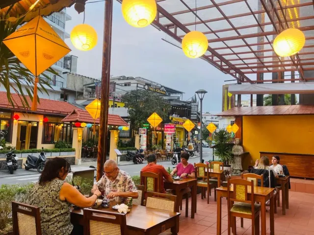 A terrace at dusk with yellow paper lanterns, wooden tables and diners facing the street