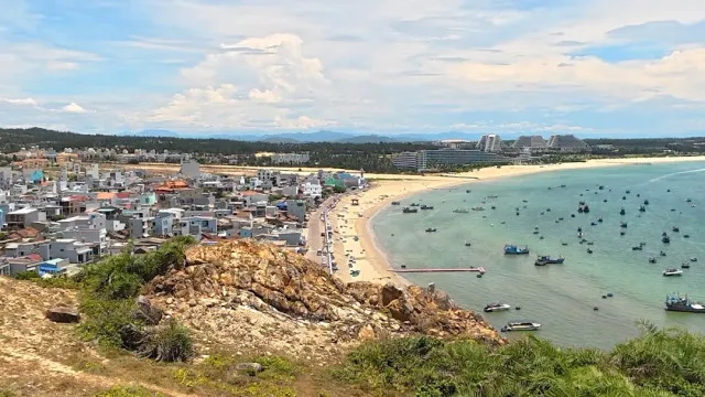 A view from a rocky hill over a dense town beside a curving beach and a bay full of boats