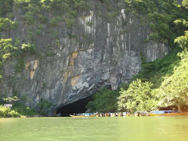 A vast cave mouth in a limestone cliff above a green river, with tour boats at the entrance