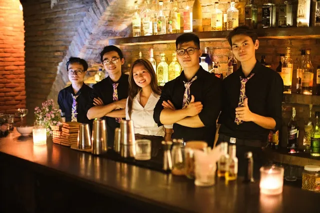 A team of bartenders in ties stands behind a counter facing a brick wall and bottle shelves
