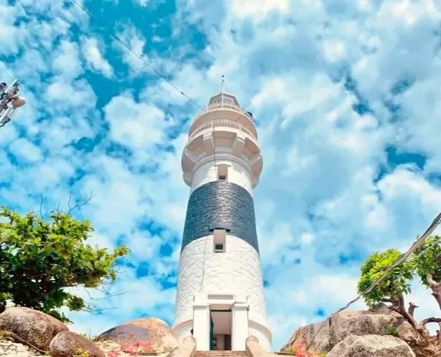 A white lighthouse with a dark band seen from below, rocks and shrubs at its base