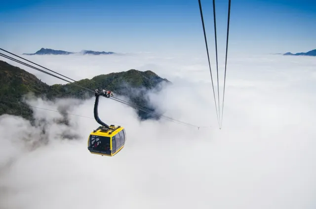 A yellow cable car cabin hanging above a sea of clouds, a green ridge showing at left