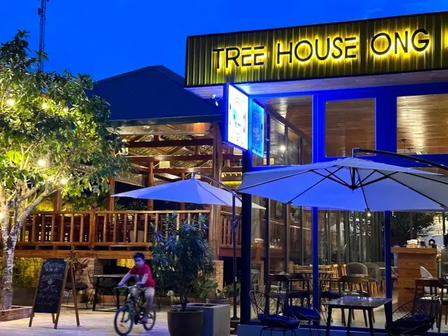 A blue hour facade with yellow neon letters, white parasols and a child riding a bicycle