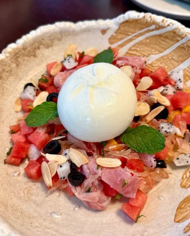 A ball of white cheese on a bed of diced watermelon, sliced almonds, black olives and mint leaves
