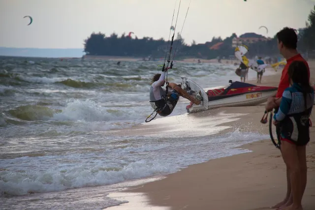 A kitesurfer with gear on a wave-swept beach, more surfers behind