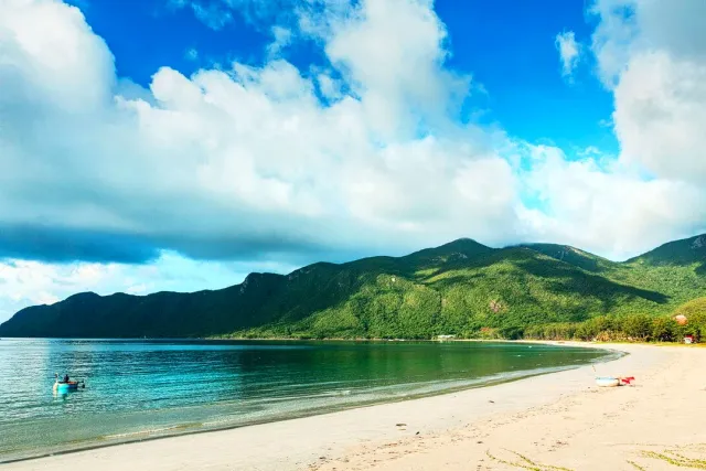 A curving bay with turquoise shallows, a green mountain ridge and big white clouds