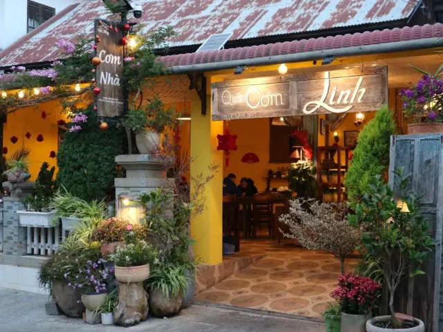 A warmly lit evening storefront with wooden signs, a yellow wall and many flowering pots at the entrance