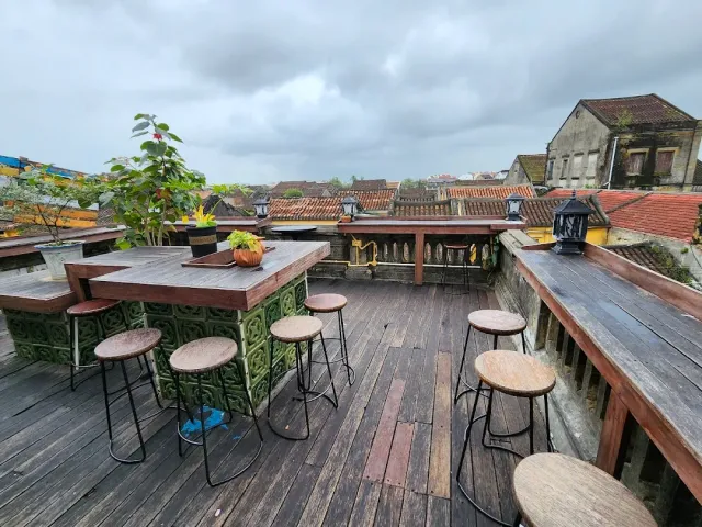 A rooftop deck with a high table and stools, a wooden railing and a view of old tiled roofs