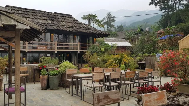 A wooden building with a wooden shingle roof, a stone terrace with chairs, and misty green hills