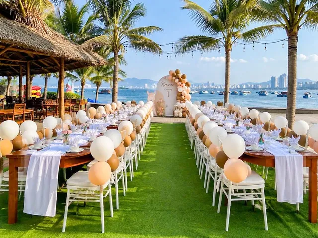 Long tables set with balloons on a lawn, facing a bay and palm trees