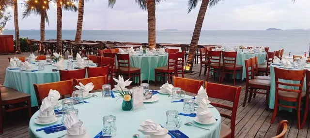 A beachfront terrace with round tables in mint cloths, palms and the sea behind