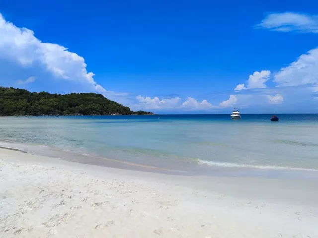 White sand and shallow turquoise water, a yacht and a boat anchored and a green headland on the left