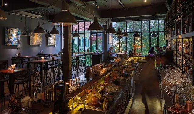 A dim bar space with a long counter, hanging lamps and tall windows onto greenery