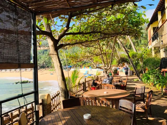 A shaded terrace with bamboo blinds, round wooden tables and a view of a sandy beach and sea