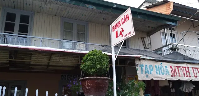 A white sign on a pole in the street, an old wooden house with a balcony and a hanging planter behind
