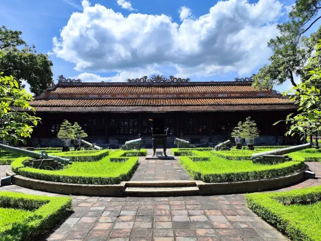 A long wooden pavilion with a tiled roof, a garden of clipped hedges and a brick path