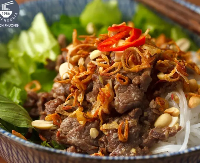 A bowl of rice noodles with beef strips, fried shallots, peanuts and red chili