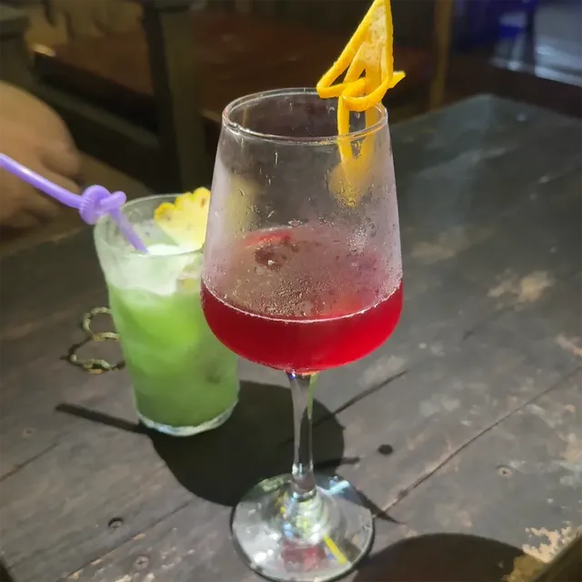 A red cocktail in a wine glass beside a green drink with pineapple, on a wooden table