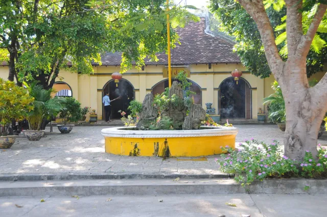 A paved courtyard with a round stone basin facing a yellow building with three arches
