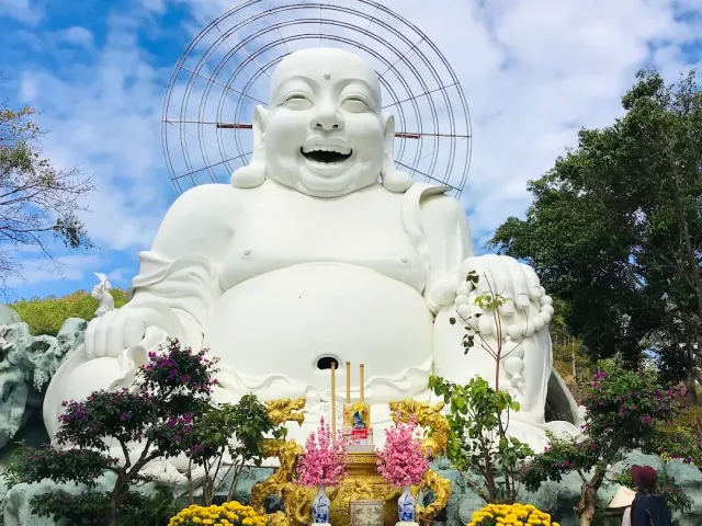 A huge white laughing Buddha statue against a blue sky, with flower beds in front of it