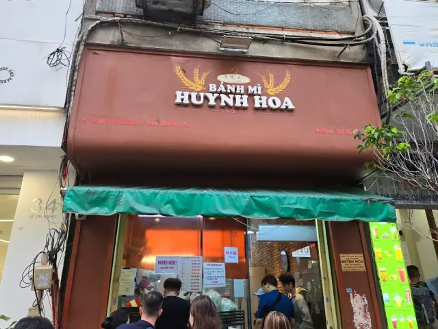 A brown shopfront with a sign and a green awning, a crowd waiting at the entrance