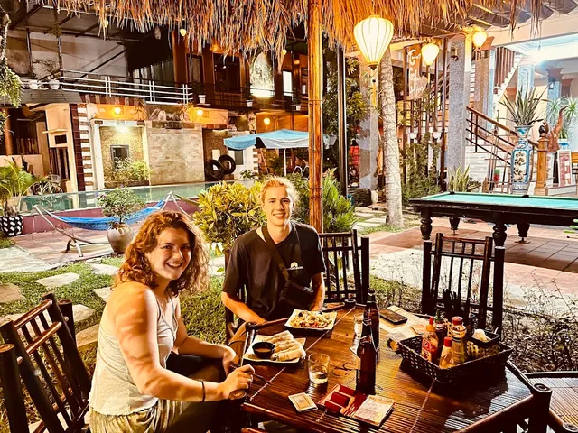 A couple dining in a lit courtyard in the evening, with a pool, a hammock and a billiard table behind
