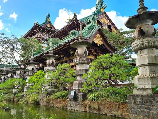 An ornate hall with a green and gold roof, a row of stone lanterns and a pond in front