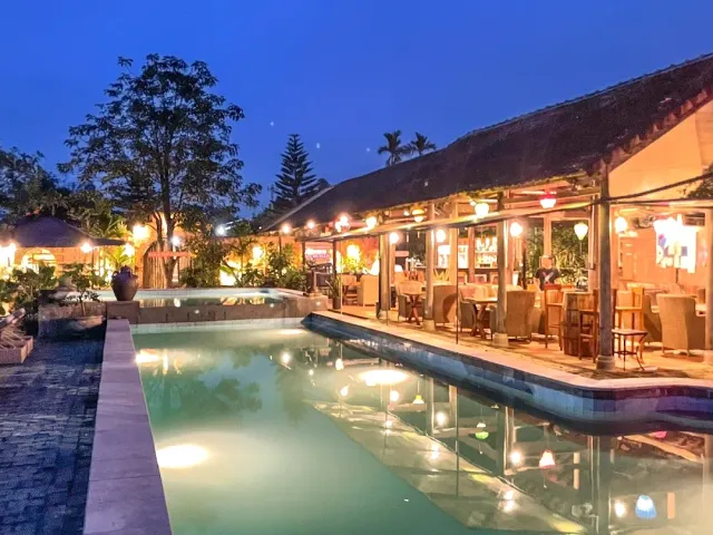 A lit pool at dusk, beside it an open thatched roof restaurant with warm lamps