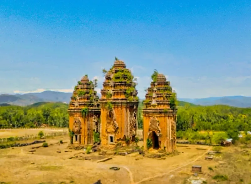 The three brick towers of Duong Long on a dry plain, with plants growing from the tops and hills behind