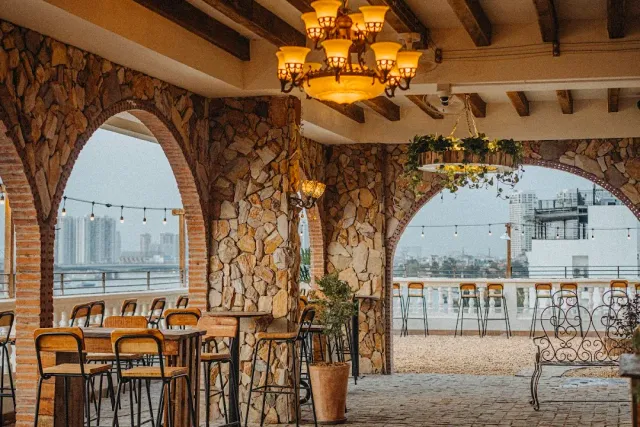 A covered rooftop with stone arches, chandeliers and high bar stools facing the city skyline