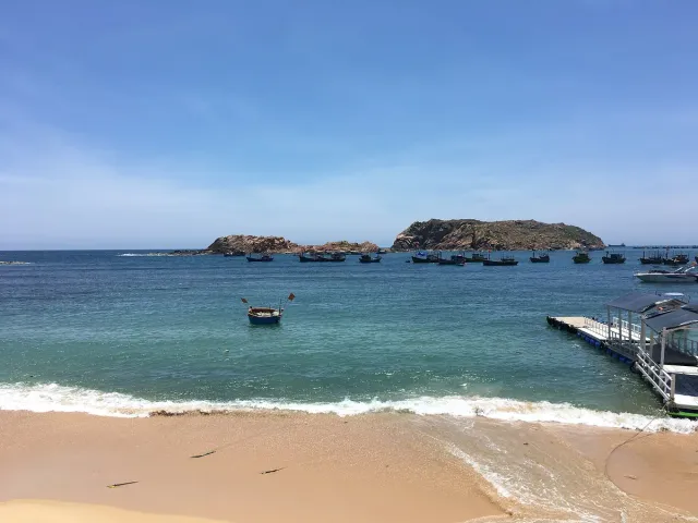 A beach with a breaking wave, a blue bay with many fishing boats and a rock mass in the water