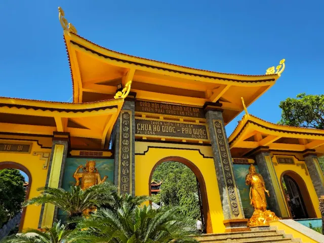 A yellow gate with curved roofs, gilded guardian statues in the arches and green plants at its foot