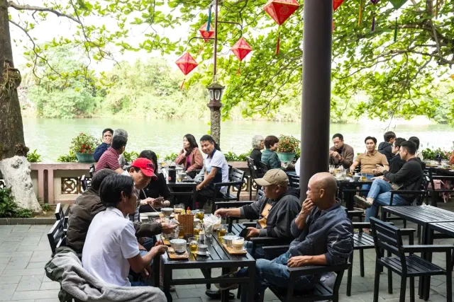 Outdoor seating under trees on a riverbank, hanging paper lanterns and diners with glasses of beer