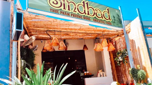 A front with a green sign, a bamboo awning, hanging conical lampshades and plants at the entrance