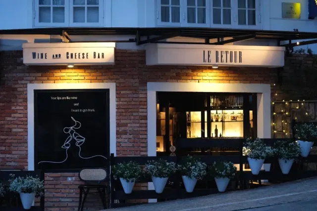 A brick facade in the evening with two lit signs and a row of white flowering planters