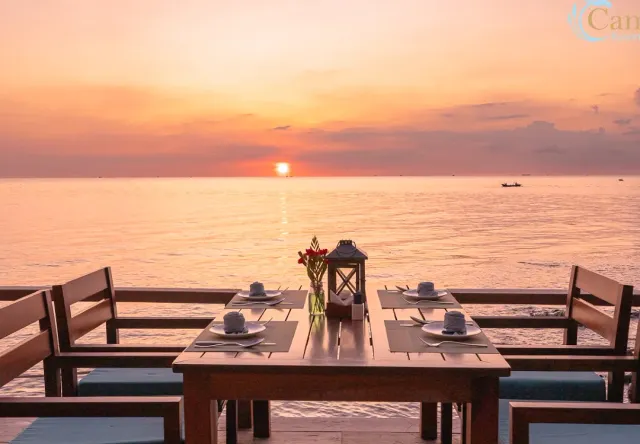 A table set on a wooden deck facing the sea at sunset, with a lantern and flowers at its center