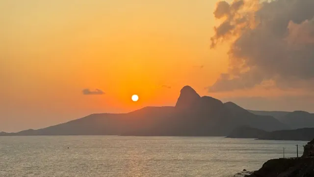 An orange sunset, the sun low over the silhouette of a pointed mountain across calm water