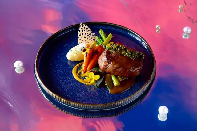 A plated meat dish on a dark blue plate over a surface in pink and blue tones