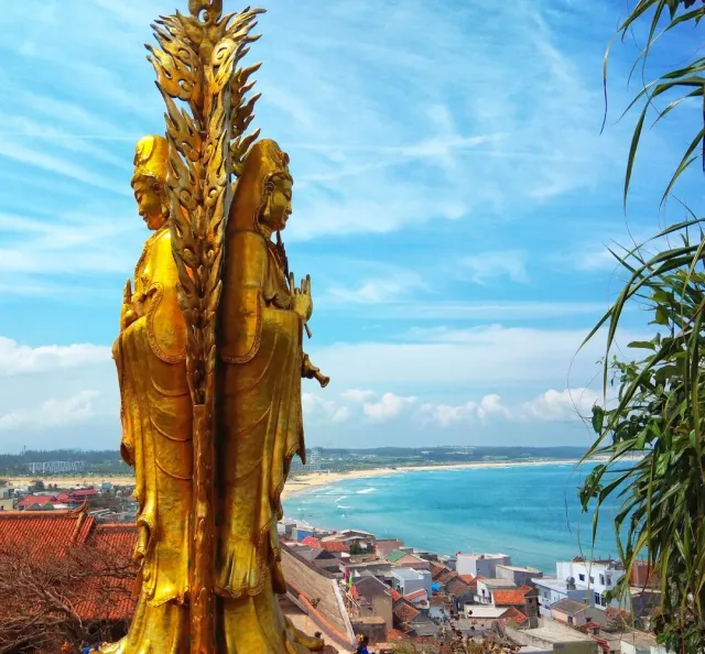 A gilded many faced statue in the foreground, with a turquoise bay and village rooftops behind it
