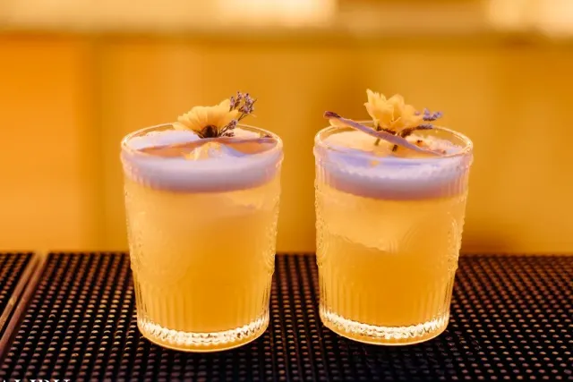 Two cocktail glasses with foam and dried flower garnish on a bar mat in amber light